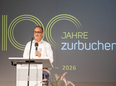 Ansprache auf der Bühne vor dem Jubiläumslogo «100 Jahre zurbuchen»